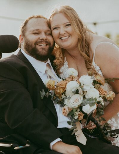 A couple smiles together outdoors. The man is in a wheelchair, wearing a suit, and the woman in a wedding dress holds a bouquet of flowers.