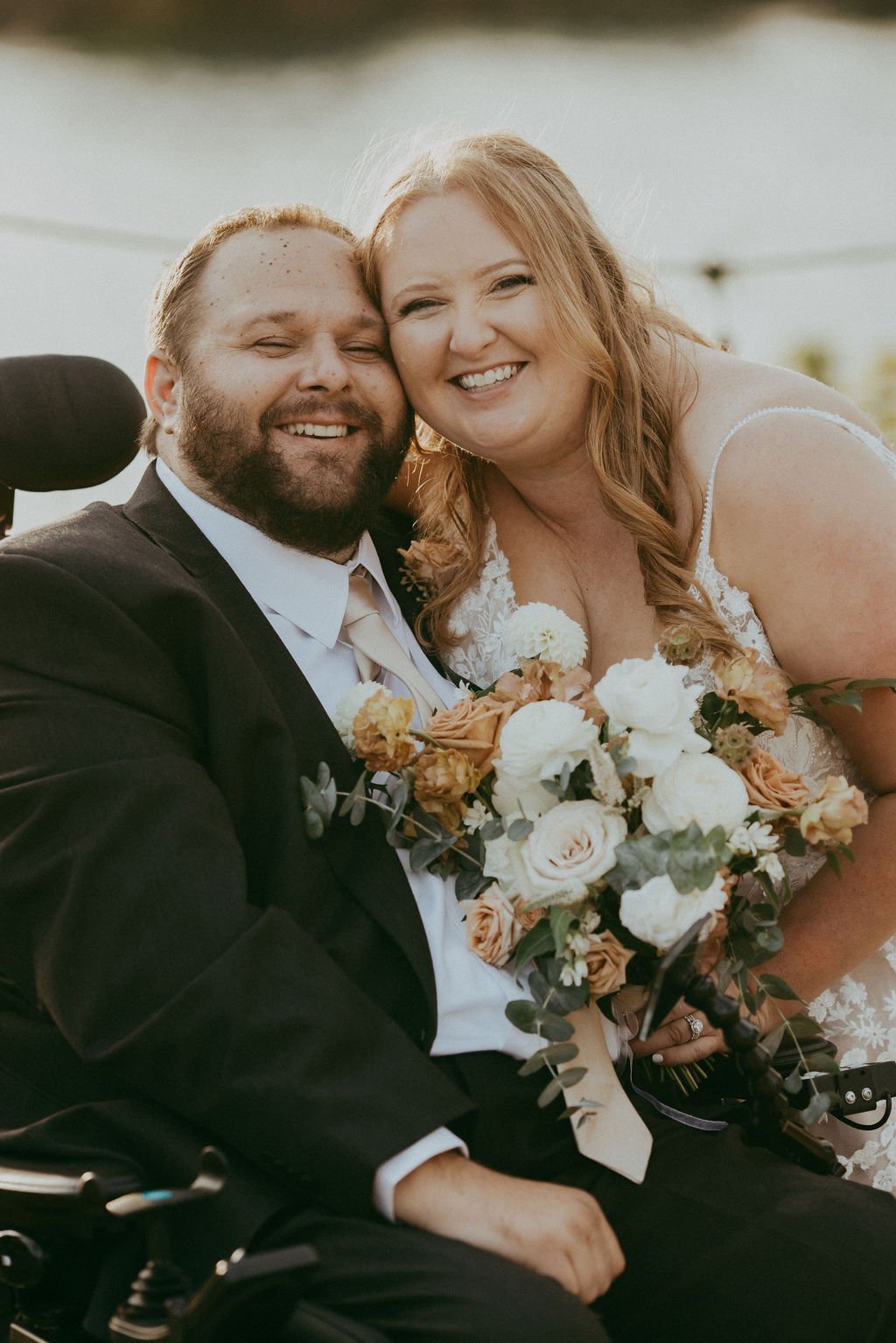 A couple smiles together outdoors. The man is in a wheelchair, wearing a suit, and the woman in a wedding dress holds a bouquet of flowers.