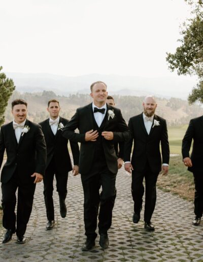 A group of six men in tuxedos walk outdoors on a stone path, surrounded by trees and greenery.