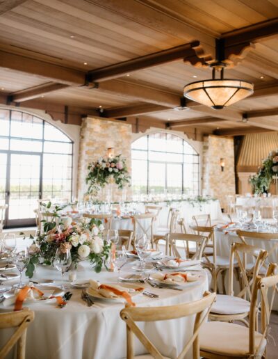 Elegant wedding reception setup with round tables, white linens, floral centerpieces, and wooden chairs. Large windows and a decorative ceiling create a warm atmosphere.