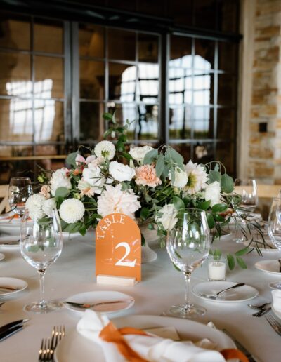 Elegant dining table set for an event with a floral centerpiece, numbered table sign, white napkins, and orange ribbon accents.