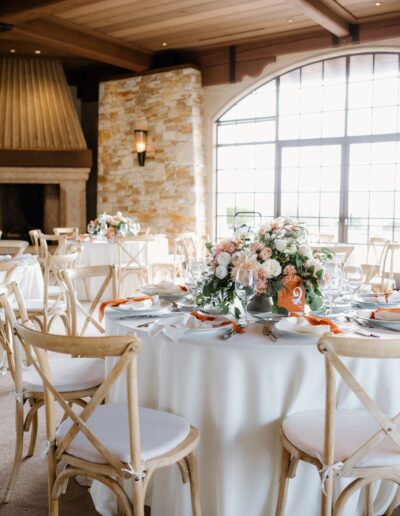 A decorated wedding reception table with a floral centerpiece, white tablecloth, and wooden chairs in a bright room with large windows and stone walls.