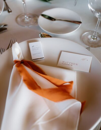 A table setting with a white plate, an orange and white napkin, a matchbox, and a place card with the name "Matthew Landrum.