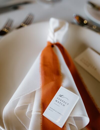 Close-up of an orange and white napkin tied with a matchbox on a plate, next to cutlery and a blurred card.