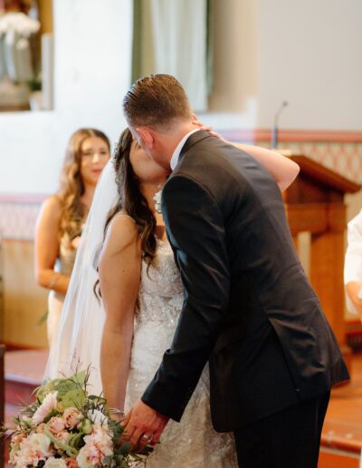 A bride and groom share a kiss at their wedding ceremony. The bride holds a bouquet of flowers. A bridesmaid stands in the background.