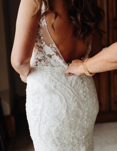 A woman wearing a lace wedding dress is being assisted by another person adjusting the dress from behind.