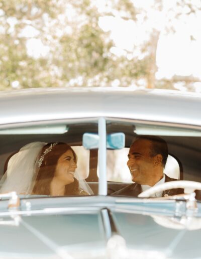 A bride and a man smiling at each other inside a vintage car.