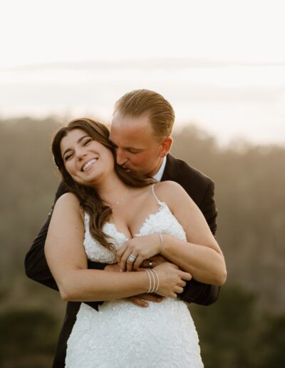 A couple embraces outdoors, with the woman in a white dress and the man in a dark suit, set against a blurred natural background.