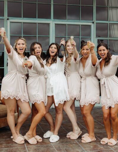 A group of six women in matching robes, one in white and the others in beige, holding drinks and smiling in a celebratory pose outdoors.