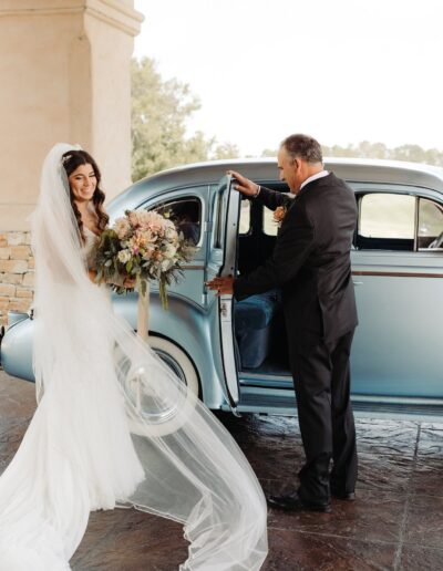 Bride in a white gown stands beside a classic blue car, holding a bouquet. A man in a suit opens the car door. Trees and stone pillars are in the background.