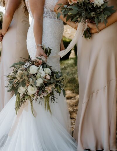 A bridal party stands outdoors holding bouquets, with the bride in a white gown and bridesmaids in beige dresses.