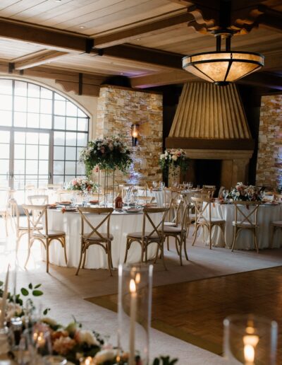 Elegant wedding reception hall with round tables, white tablecloths, floral centerpieces, and a large stone fireplace under a wooden ceiling.