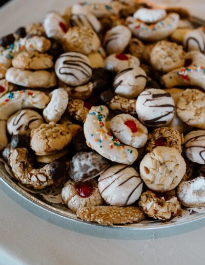 A platter filled with a variety of cookies, including ones with chocolate drizzle, powdered sugar, and colorful sprinkles, displayed on a white tablecloth.