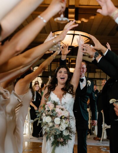 Bride and groom walk joyfully through a tunnel formed by guests' raised arms at a wedding reception. Bride holds a floral bouquet.