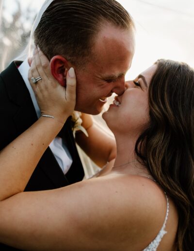 A bride and groom embrace and smile under a sheer veil outdoors, with the bride touching the groom's face.