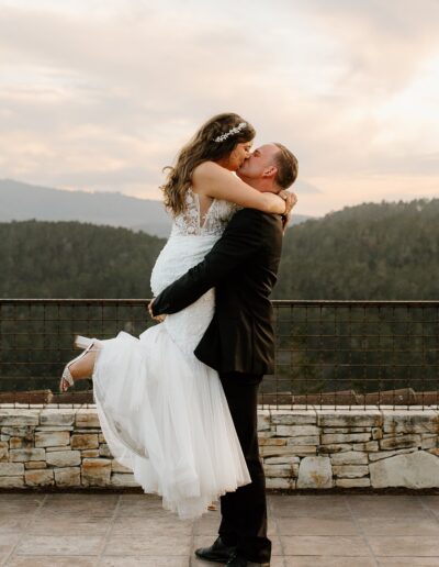 A groom lifts a bride in a white dress while they kiss, set against a scenic mountain backdrop with a stone railing.