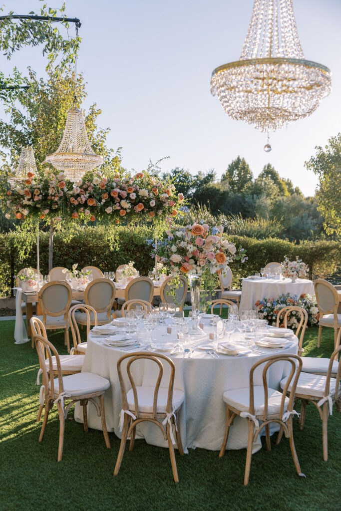 Outdoor wedding setup with round tables decorated with flowers and white linens. Crystal chandeliers hang above, and greenery surrounds the area.