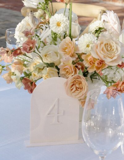 Elegant floral centerpiece with roses and assorted flowers on a table, accompanied by a table number card displaying the number "4," with empty wine glasses nearby.