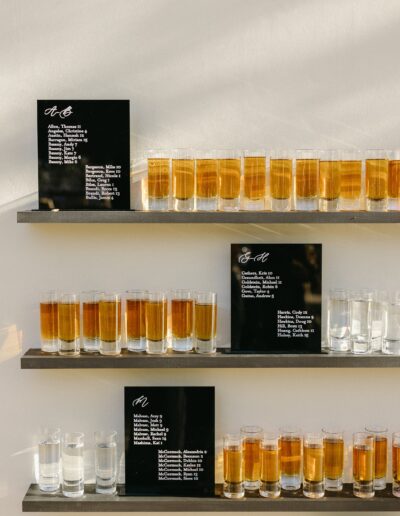 Three shelves display rows of filled shot glasses. Each shelf includes a black sign with a list of ingredient names.