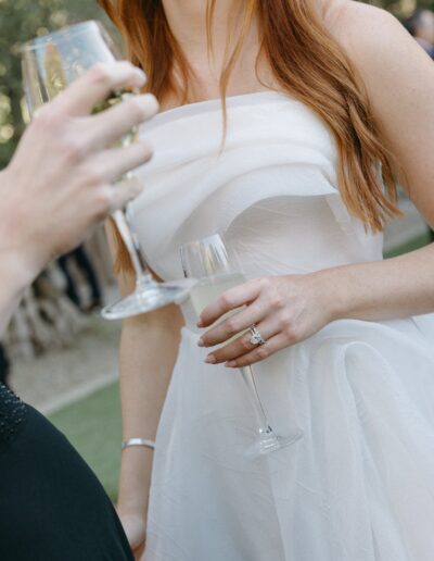 Two people holding champagne flutes, one wearing a white dress and the other in black attire, possibly at an outdoor gathering.