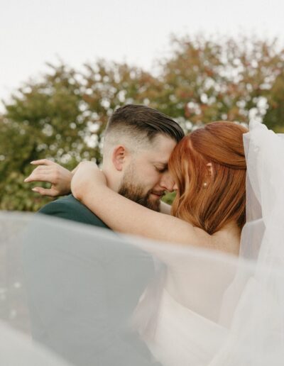 A couple embraces closely outdoors, surrounded by greenery. The bride's red hair and the groom's dark hair are visible, along with a flowing veil.