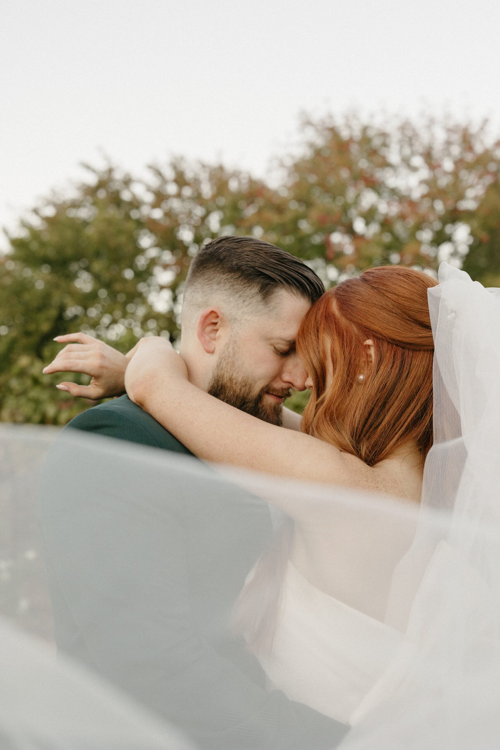 A couple embraces closely outdoors, surrounded by greenery. The bride's red hair and the groom's dark hair are visible, along with a flowing veil.