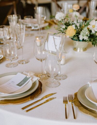 Elegant table setting with neatly arranged plates, gold cutlery, wine glasses, and a centerpiece with white and yellow flowers on a white tablecloth.