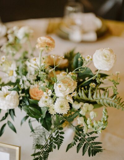 A floral arrangement with white and peach flowers, greenery, and ferns on a table.