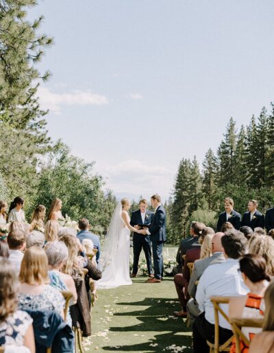 A couple stands before an officiant during an outdoor wedding ceremony, surrounded by guests seated in rows, with a backdrop of trees and a clear sky.