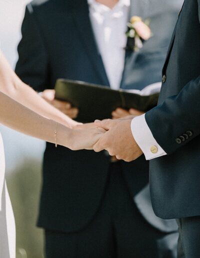 A bride and groom holding hands during a wedding ceremony, with an officiant in the background holding a book.