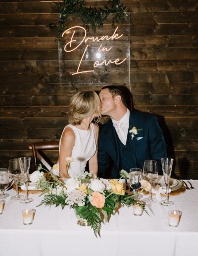 A couple kisses at a wedding reception table with a "Drunk in Love" sign behind them.