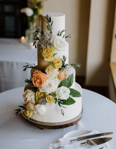 Three-tiered white wedding cake adorned with white, yellow, and peach roses, placed on a round table with two cake knives on a napkin beside it.