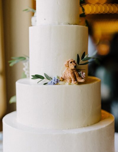 A tiered white cake with a small dog figurine and greenery decoration on one layer.