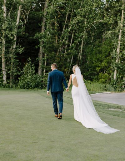 Bride and groom walk hand in hand on a grassy field, surrounded by tall trees. The bride wears a long white gown with a veil, and the groom is in a blue suit.