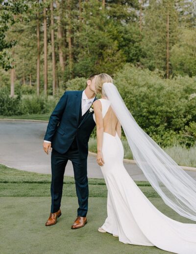 A couple in formal attire shares a kiss outdoors, with lush greenery and a path in the background. The woman wears a long, flowing veil.