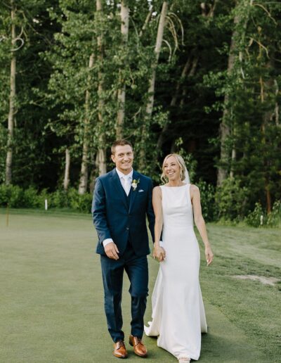 A couple in formal attire walks hand in hand on a grassy area, surrounded by tall trees.