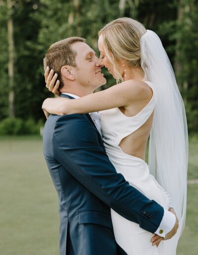 A bride and groom embrace and smile at each other outdoors, with green trees in the background. She wears a long white dress and veil, and he is in a blue suit.