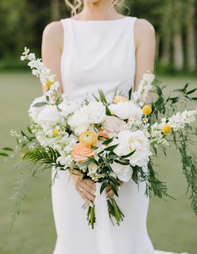 A woman in a white sleeveless dress holding a bouquet of white, yellow, and peach flowers with greenery.