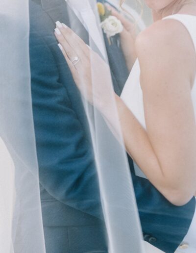 Bride and groom embracing, partially covered by a sheer veil. Bride's engagement ring and groom's suit are visible.