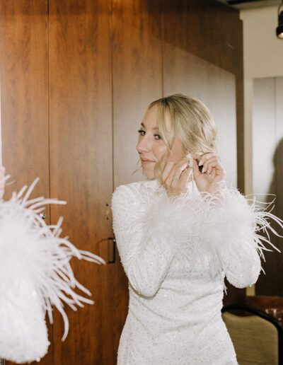 A person in a white dress with feather cuffs adjusts earrings while looking in a mirror.