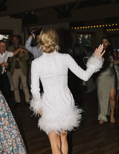 A woman in a white, feathered dress is dancing with her back to the camera. Others are dancing around her in a dimly lit room.