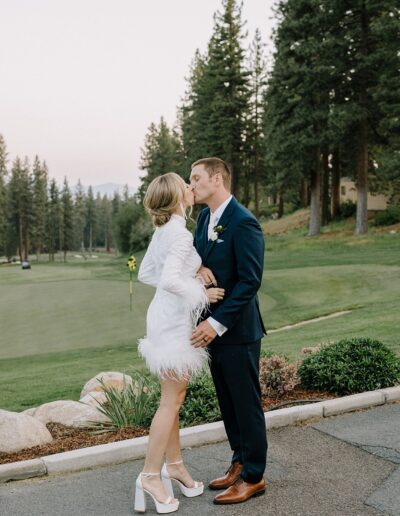 A couple kissing outdoors near a golf course, surrounded by trees. The woman is wearing a white dress with feather details, and the man is in a dark suit.