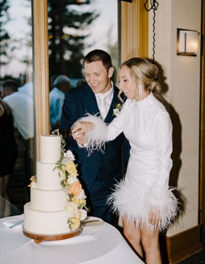 A couple in formal attire is smiling as they cut a three-tiered cake adorned with flowers at an indoor event.