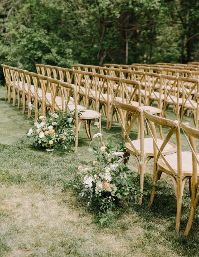 Outdoor wedding ceremony setup with rows of wooden chairs on a grassy area, surrounded by trees. Floral arrangements line the aisle.