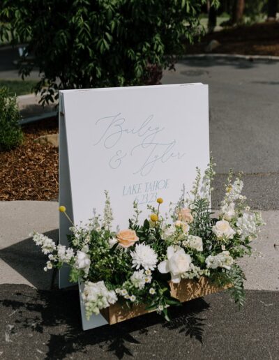 A sign with "Bailey & Tyler, Lake Tahoe, 2023" stands on a path, adorned with white and peach flowers and green leaves.