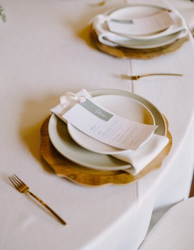 Elegant table setting with a layered white plate, napkin, and menu on a wooden charging plate, arranged on a white tablecloth with gold cutlery.