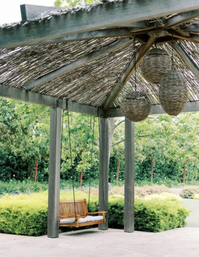 A wooden swing under a pergola with woven roof and hanging lanterns, surrounded by greenery and a stone patio.