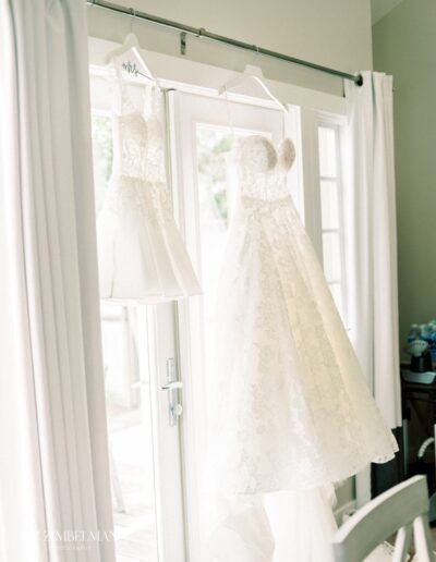 Two white lace bridal dresses hang in front of a sunny window with white curtains.