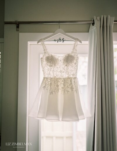 A white bridal dress with floral embroidery is hanging on a hanger labeled "mrs" near a window.