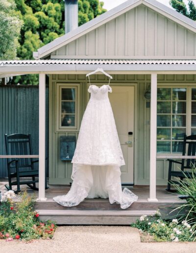 A wedding dress hangs on the porch of a small house with a white facade. Two rocking chairs are positioned on either side, and there are flowers and greenery in the foreground.